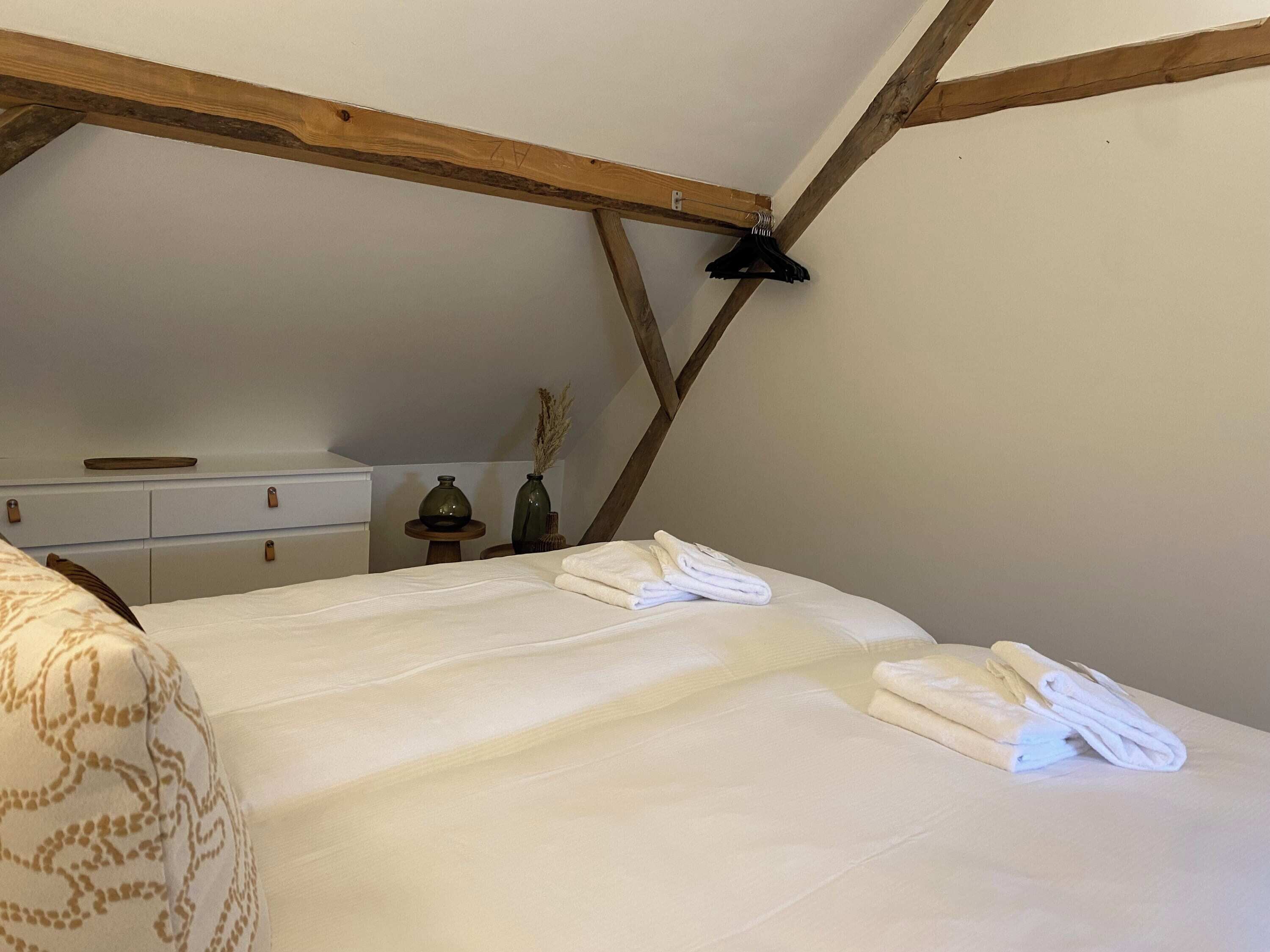 Ein Schlafzimmer mit Holzbalkendecke, einem Bett mit weißer Bettwäsche, einem Schminktisch und einem kleinen Tisch mit Vase und Kerze.
