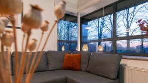 Een moderne woonkamer met een grijze modulaire bank, een groot raam met uitzicht op bomen en een vaas met gedroogde bloemen.