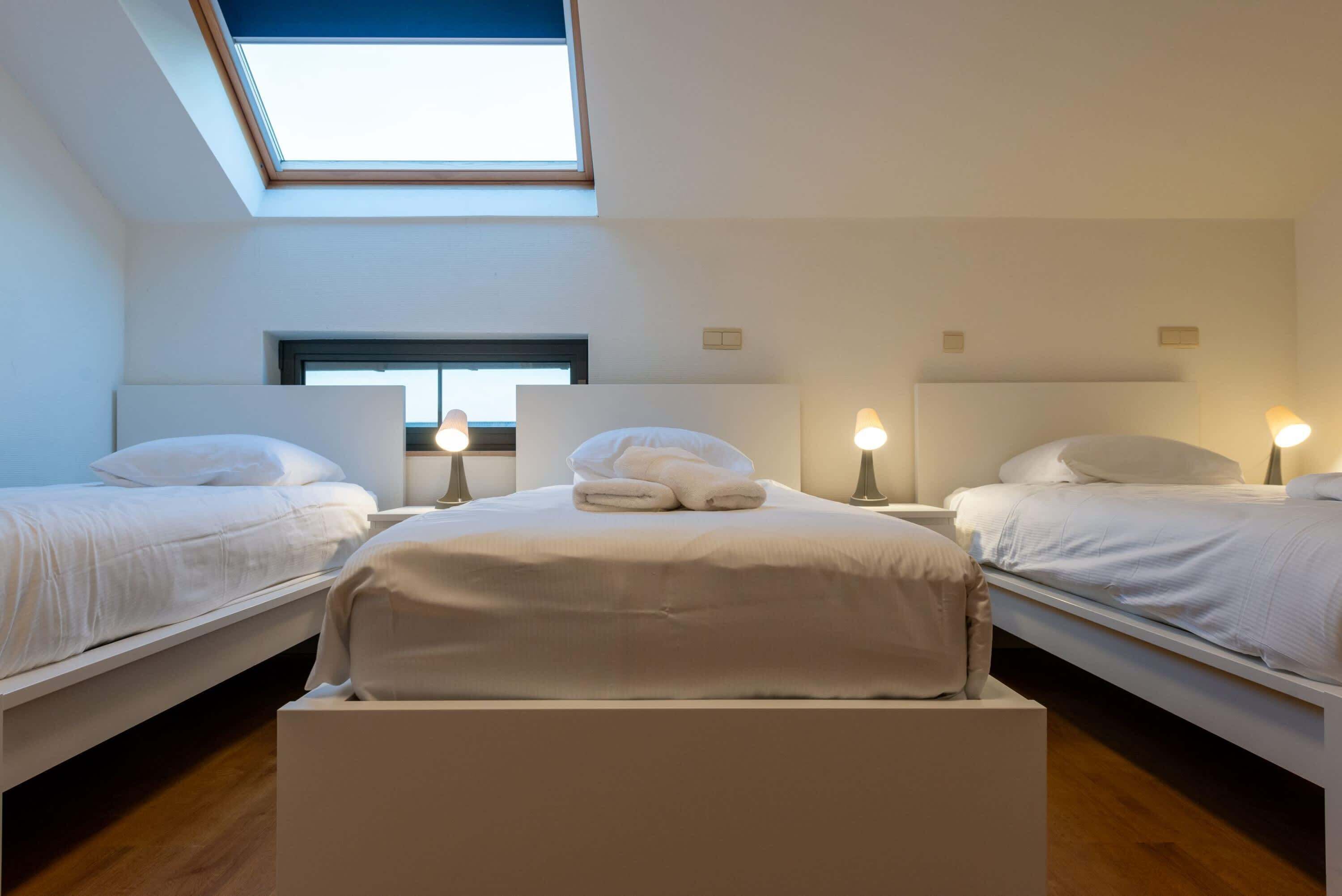 Una habitación de hotel moderna con tragaluces, dos camas individuales y una cama central, con lámparas de noche.