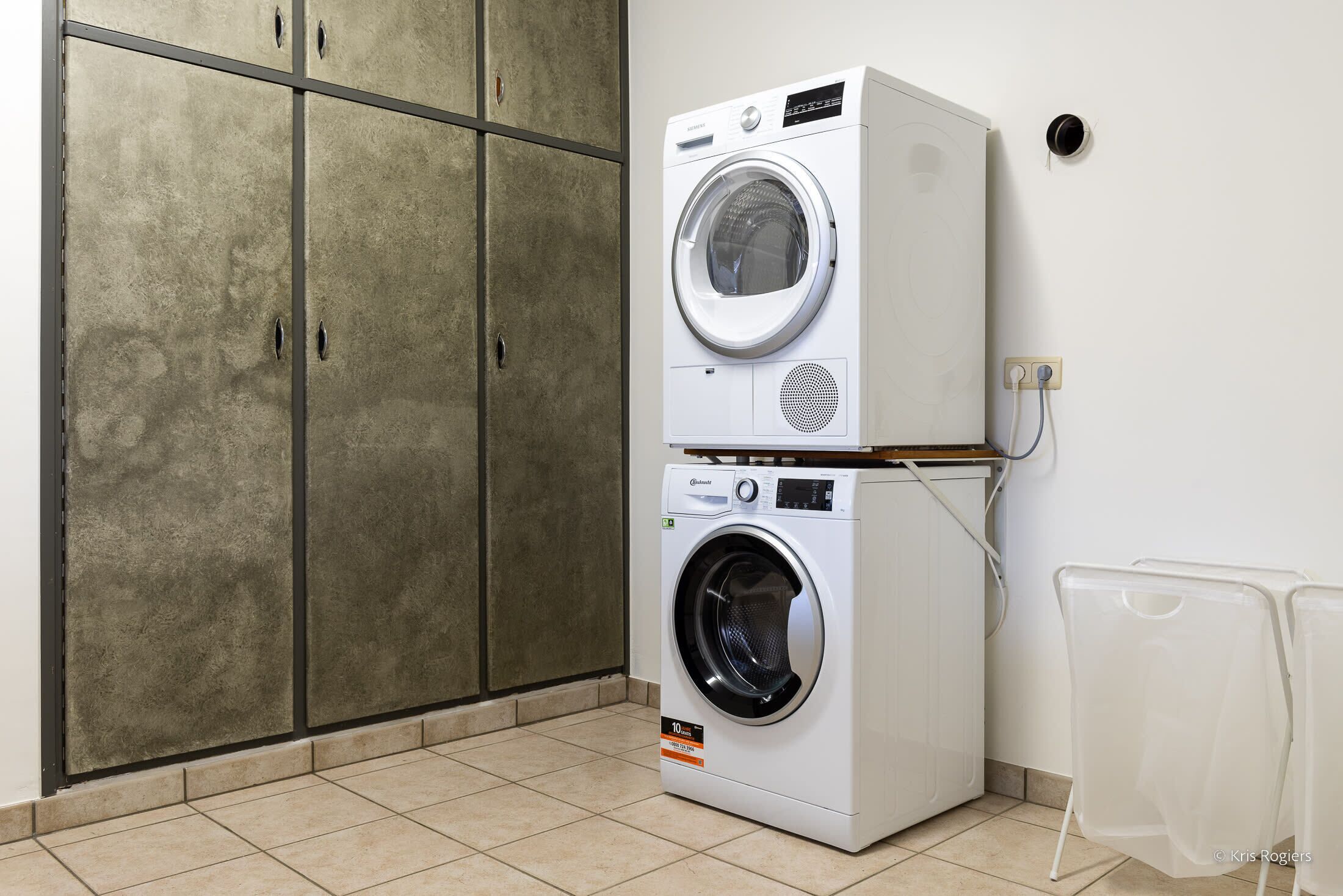 Een gestapelde wasmachine en droger in een ruimte met betegelde vloer en een grote inbouwkast.