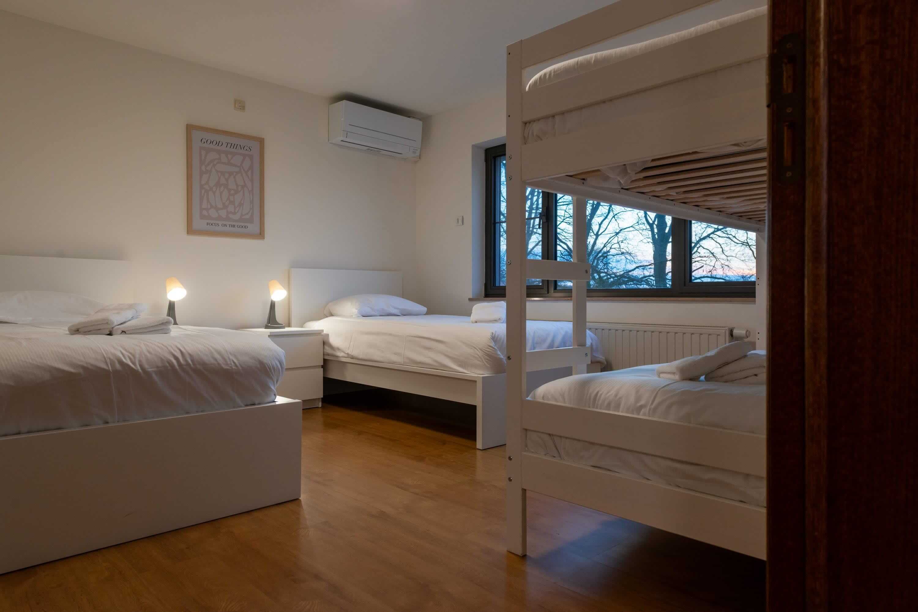 Een slaapkamer met twee stapelbedden, een nachtkastje met een lamp, een lijst met een afbeelding aan de muur, en een raam met uitzicht op bomen.