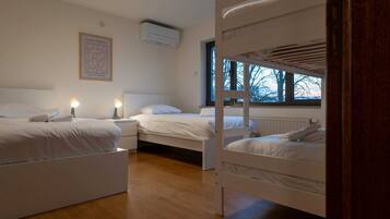 Een slaapkamer met twee stapelbedden, een nachtkastje met een lamp, een lijst met een afbeelding aan de muur, en een raam met uitzicht op bomen.