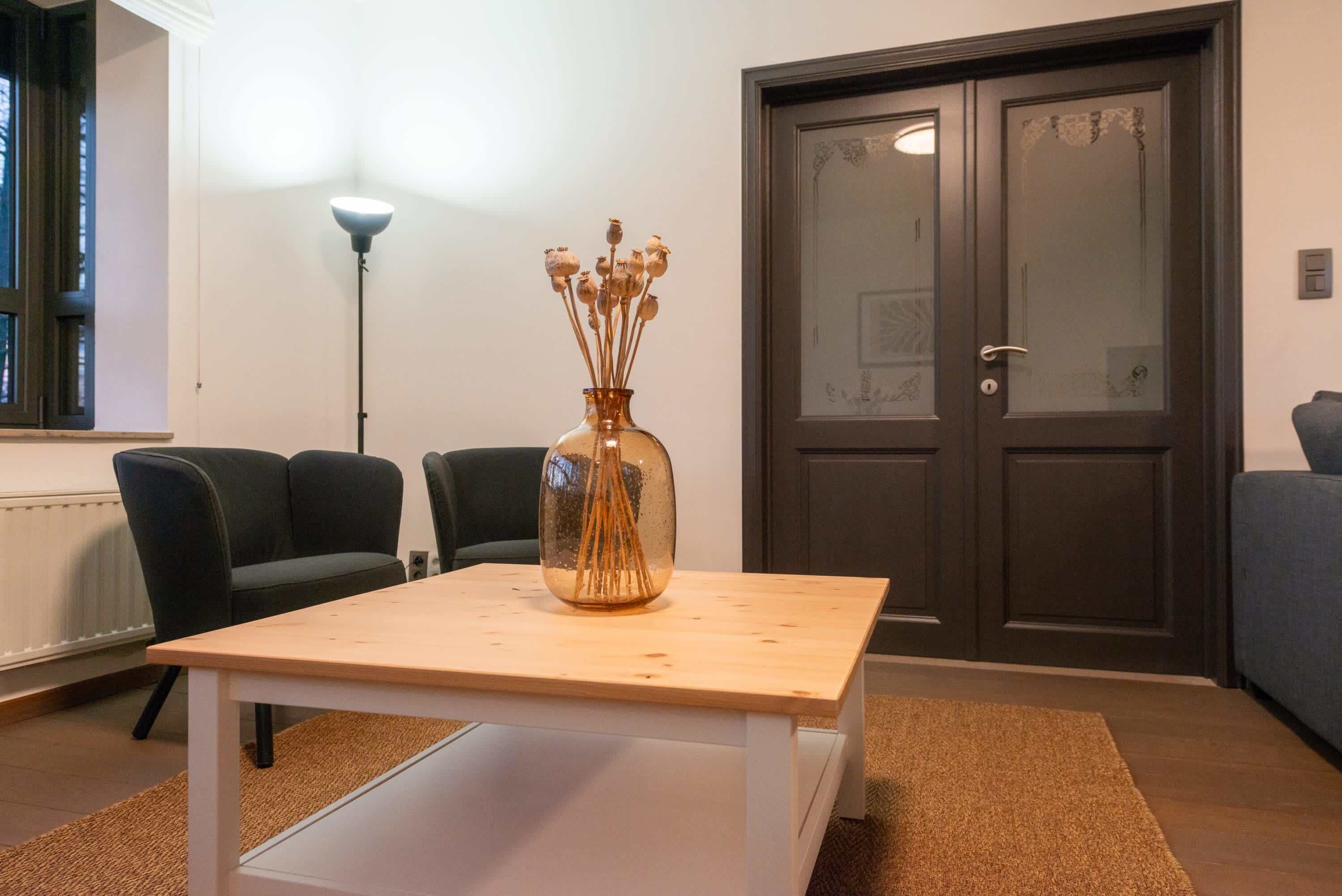 Ein Zimmer mit Holztisch, einer Vase mit getrockneten Blumen, zwei schwarzen Stühlen, einer Stehlampe und einer dunklen Holztür.