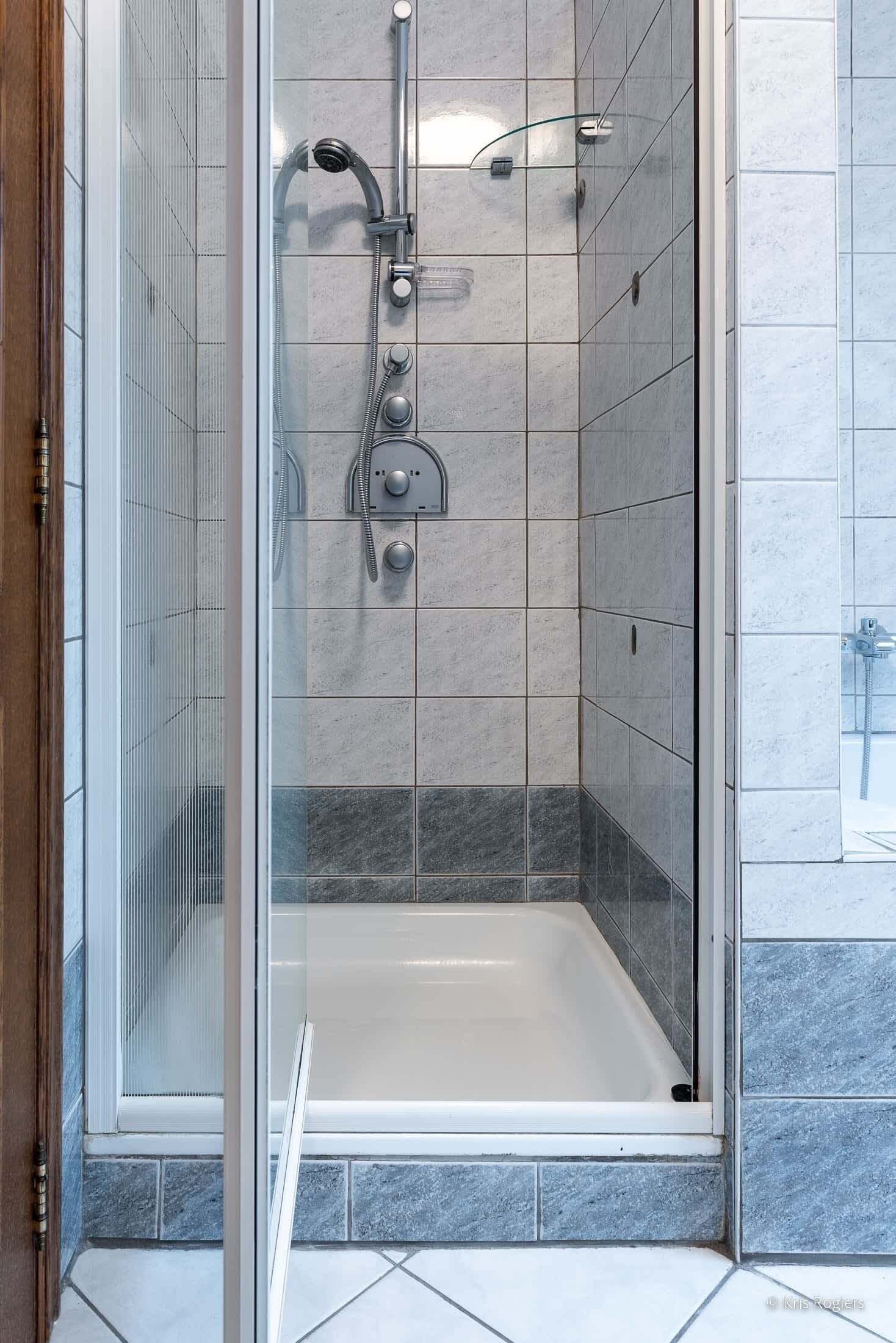 Doccia a vetro con soffione a mano, pannello comandi e vasca da bagno.