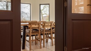 Een eetgedeelte met houten stoelen en een tafel, grote ramen en uitzicht op bomen.