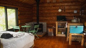 A cozy cabin with a bed, a fireplace, a small table, and a window. - Off-Grid WoodLog Cabin (Blaeberry)
