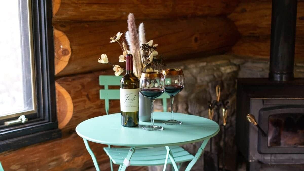 Un intérieur rustique en bois, avec une table ronde sur laquelle sont posés une bouteille et deux verres, une cheminée et un mur en pierre.