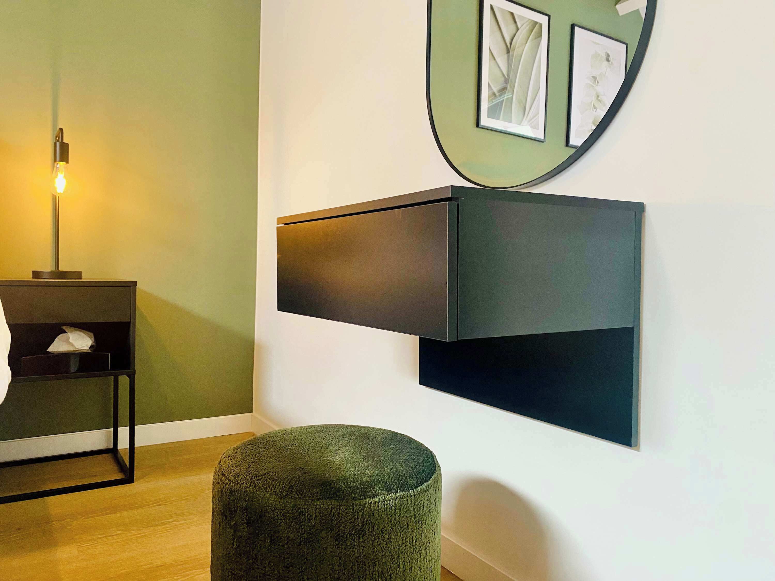 Quarto moderno com piso de madeira, parede verde, móvel de armazenamento preto, um banquinho redondo e um abajur.