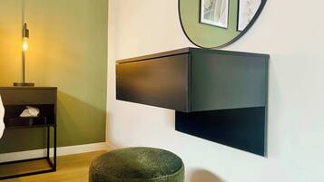 Quarto moderno com piso de madeira, parede verde, móvel de armazenamento preto, um banquinho redondo e um abajur.