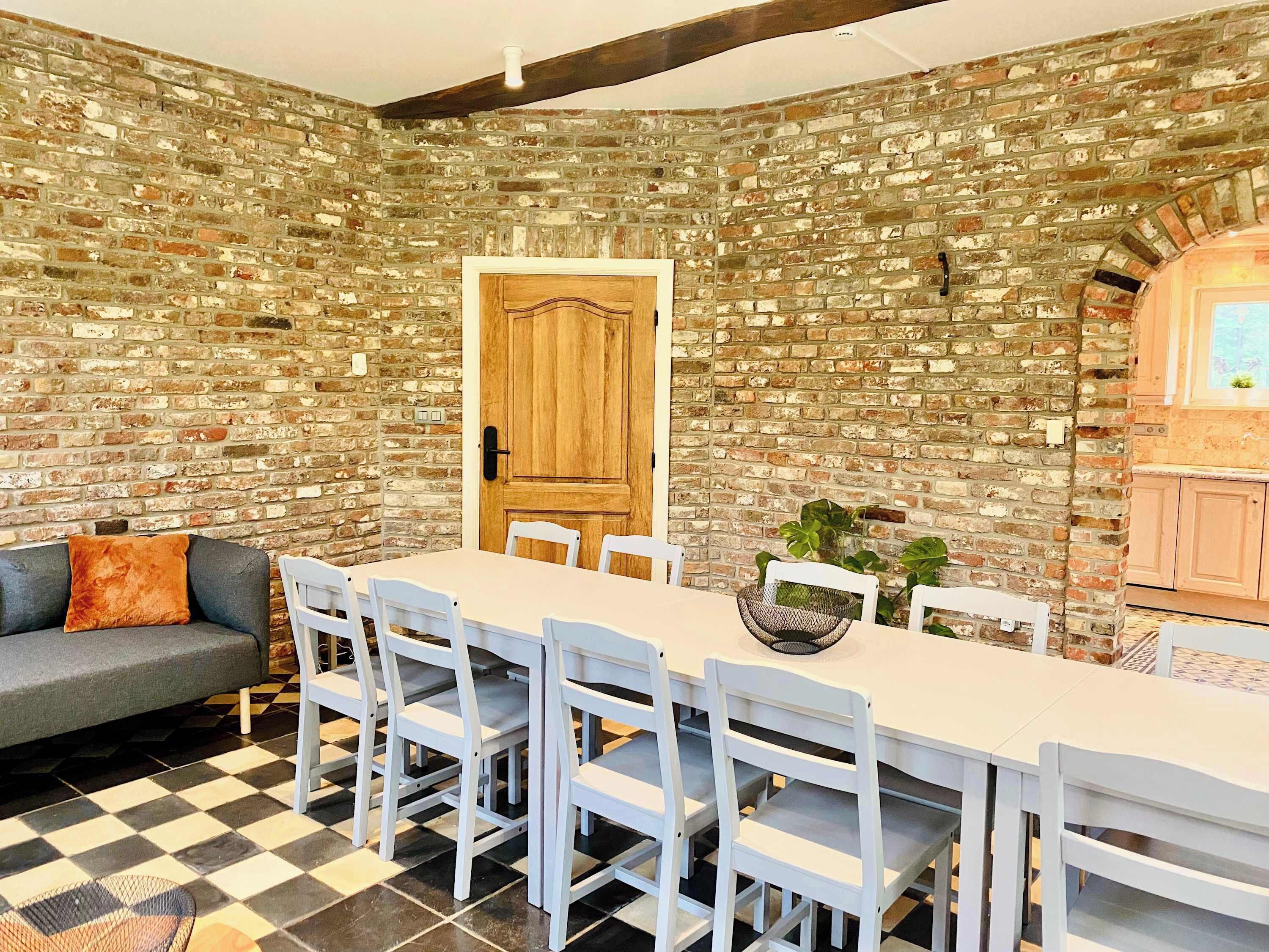 Un coin repas avec un mur en briques, une porte en bois, des chaises blanches, une table et un canapé gris agrémenté de coussins orange.