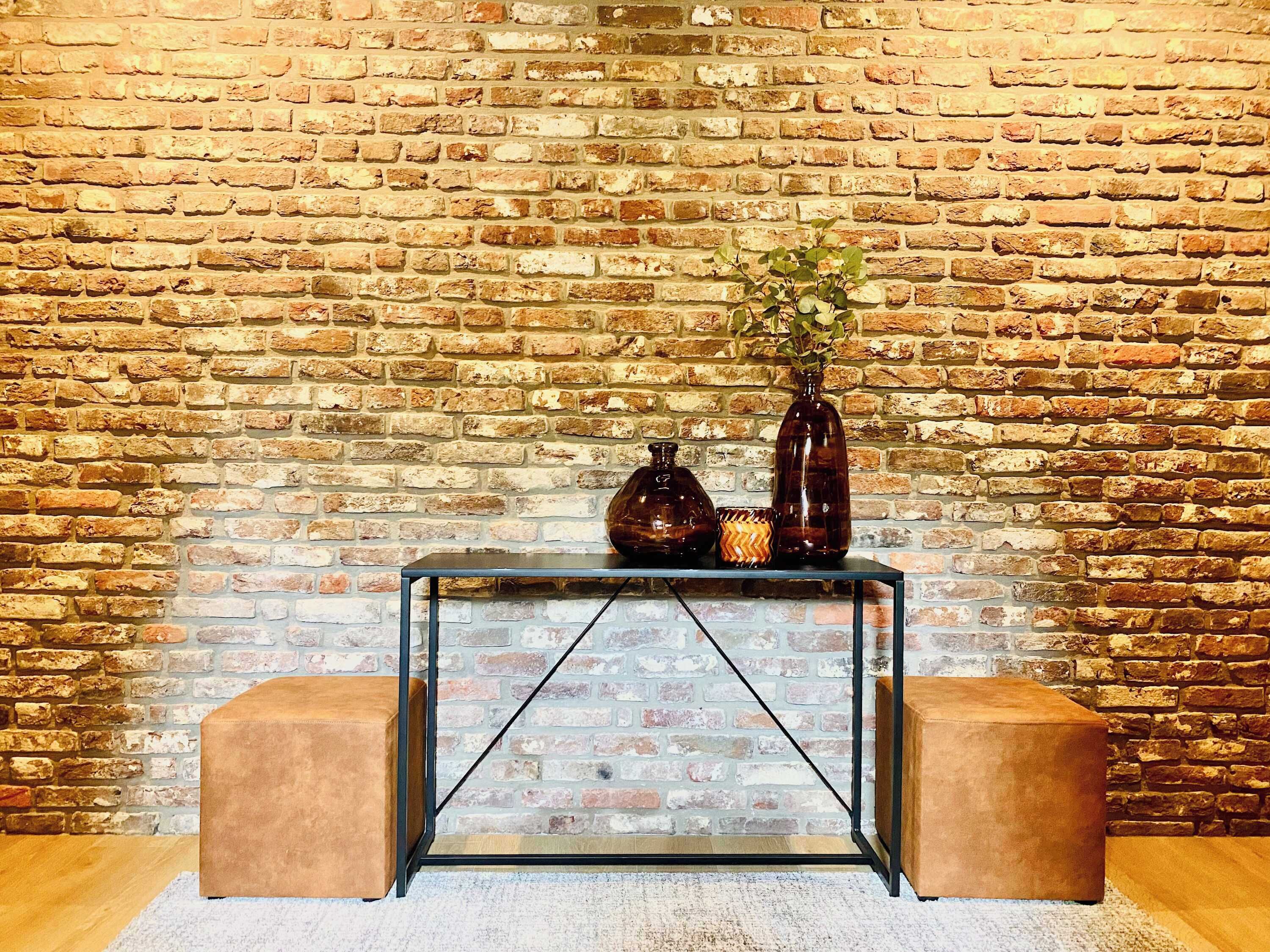 A room with a brick wall, a glass console table, two brown ottomans, and a vase with a plant.