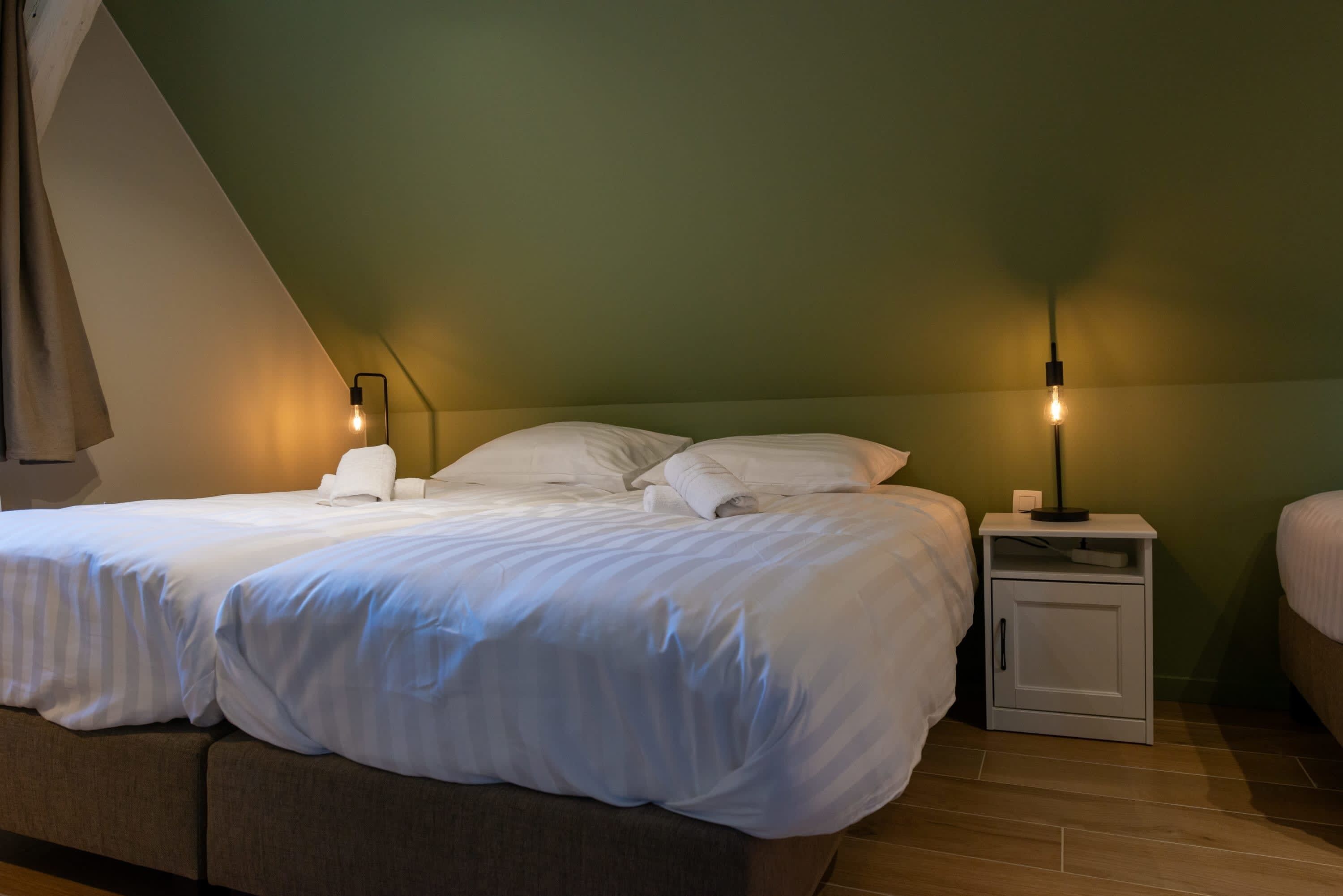 Ein ordentlich bezogenes Bett mit weißen Leinen in einem Raum mit schrägem Dach und grünen Wänden. Es gibt zwei Nachttische mit Lampen.