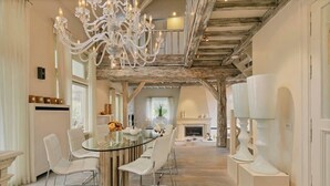 A spacious dining area with a glass table surrounded by white chairs, a grand chandelier, and exposed wooden beams.