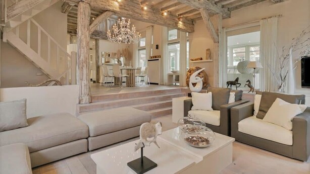 A spacious living area with a high ceiling, exposed wooden beams, a chandelier, and a staircase leading to an upper floor.