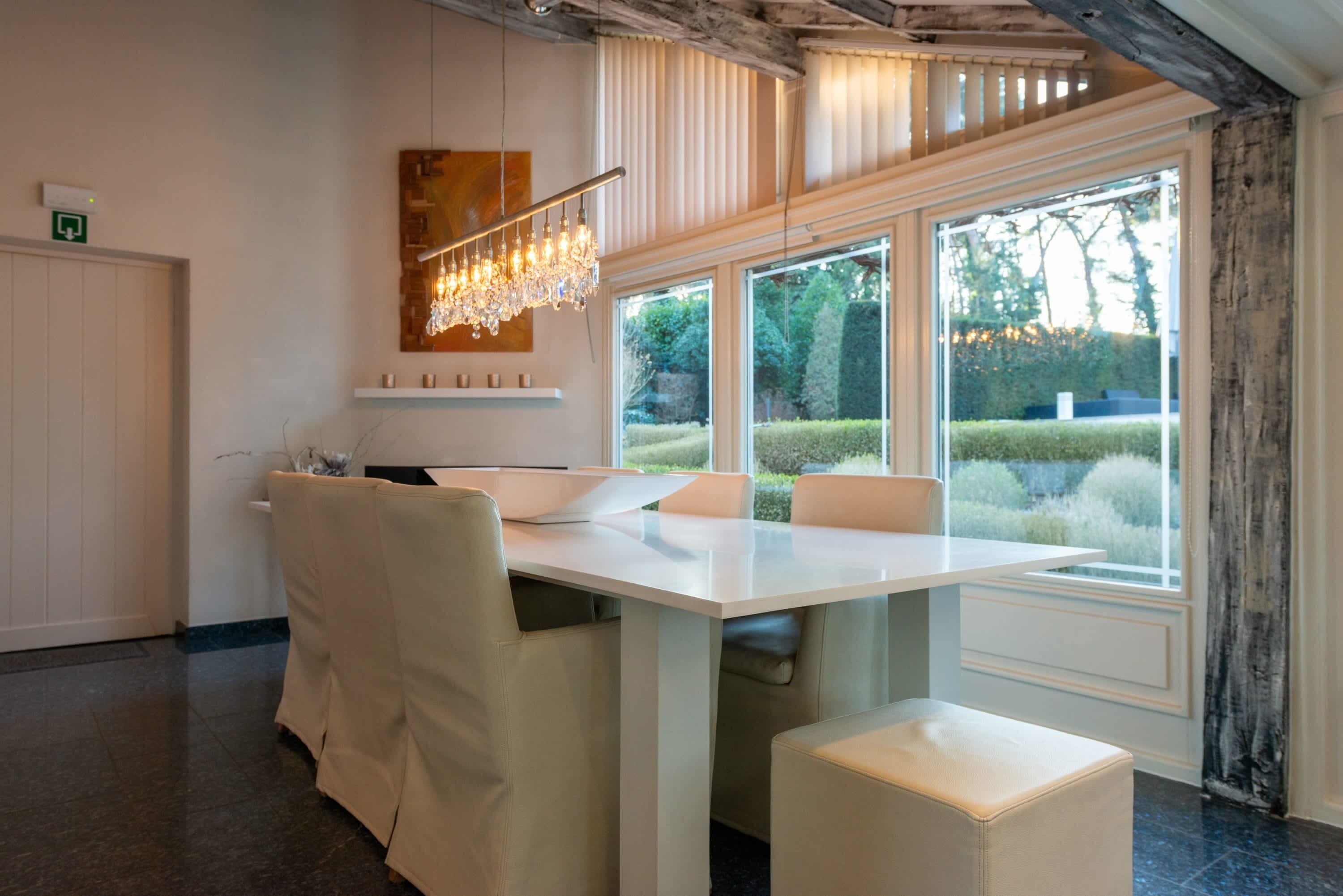 A modern dining area with a white table and chairs, a chandelier, and large windows with a view of greenery.