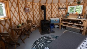 Een gezellig interieur van een joert met een houten kachel in het midden, een eethoek met stoelen, een kleine tafel en een raam met uitzicht op groen.