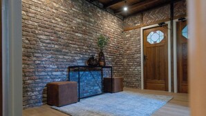 Ein Zimmer mit Backsteinwand, einer Holztür mit Glasfenster, einem kleinen Tisch mit Pflanze und zwei braunen Hocker.