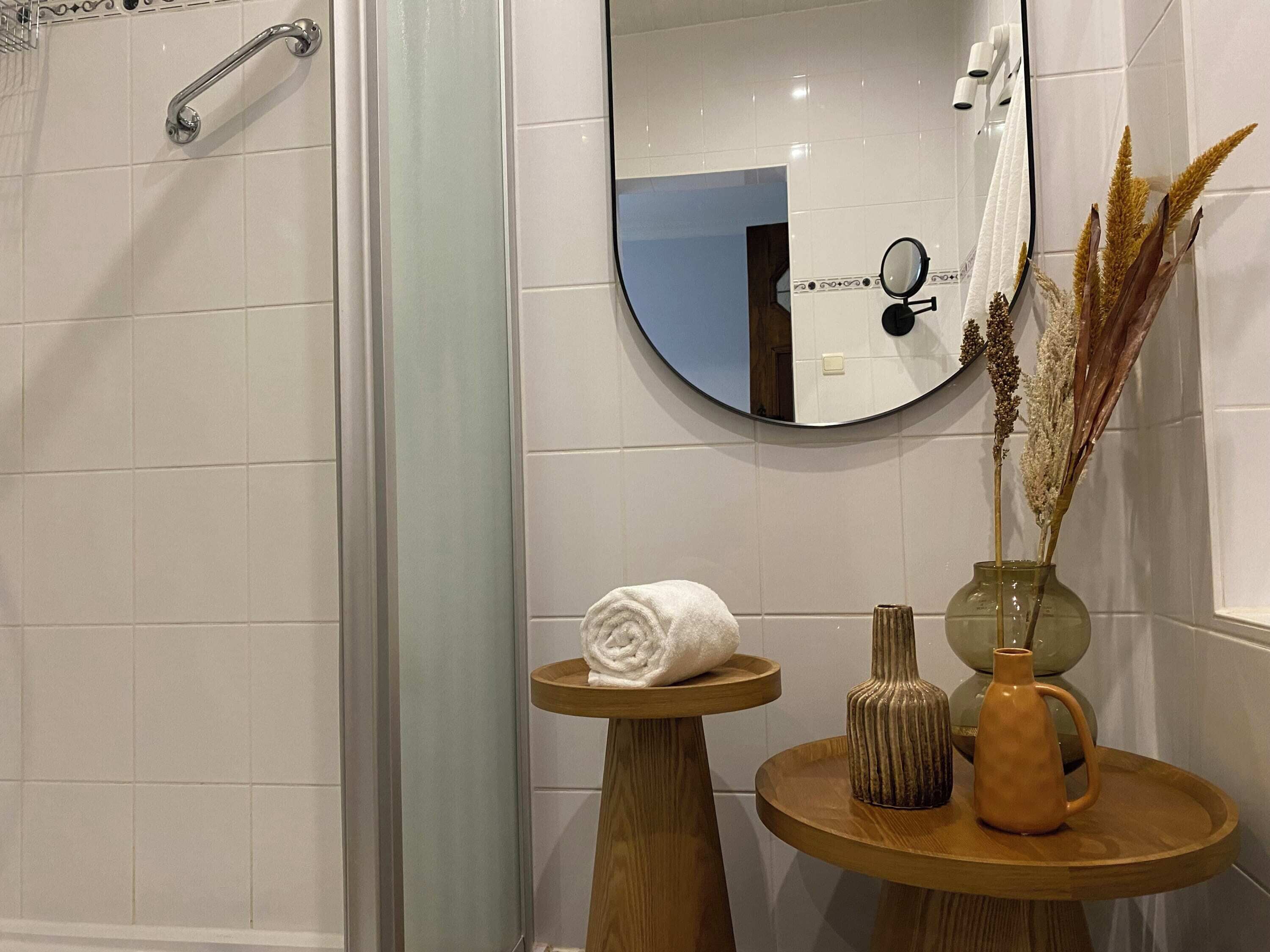 A bathroom with white tiles, a shower with a glass door, a round mirror, a towel on a wooden stool, and decorative vases.