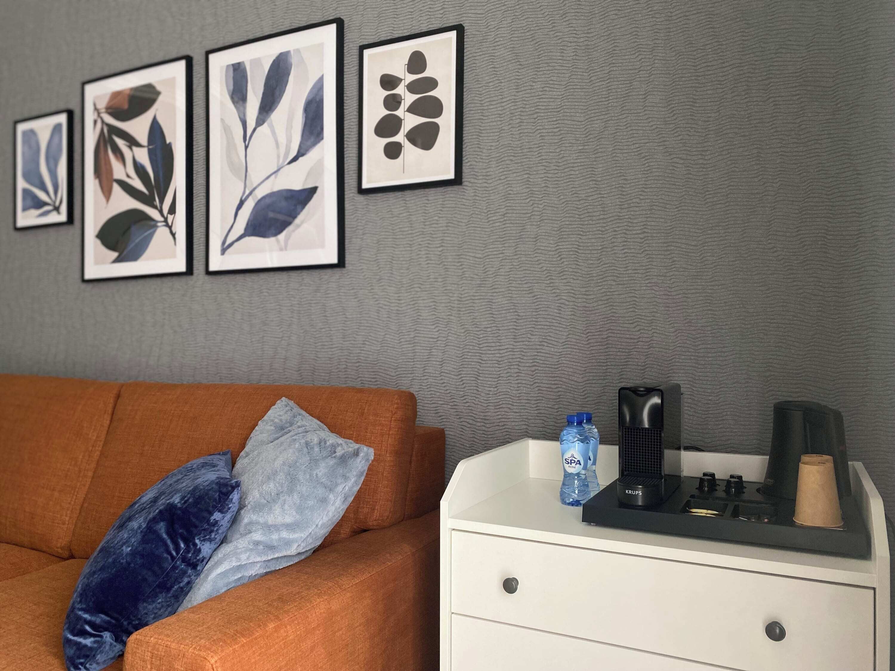 Una habitación con pared gris texturizada, una mesita de noche blanca y un sofá marrón con dos almohadas. Hay cuatro diseños de hojas enmarcados en la pared.