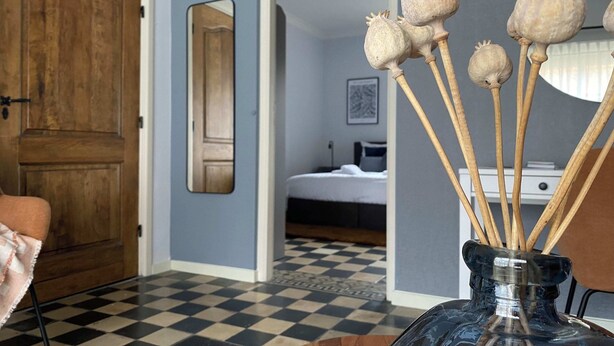 Une pièce avec un sol à carreaux, une porte en bois, un miroir et un lit recouvert d’une couverture blanche.