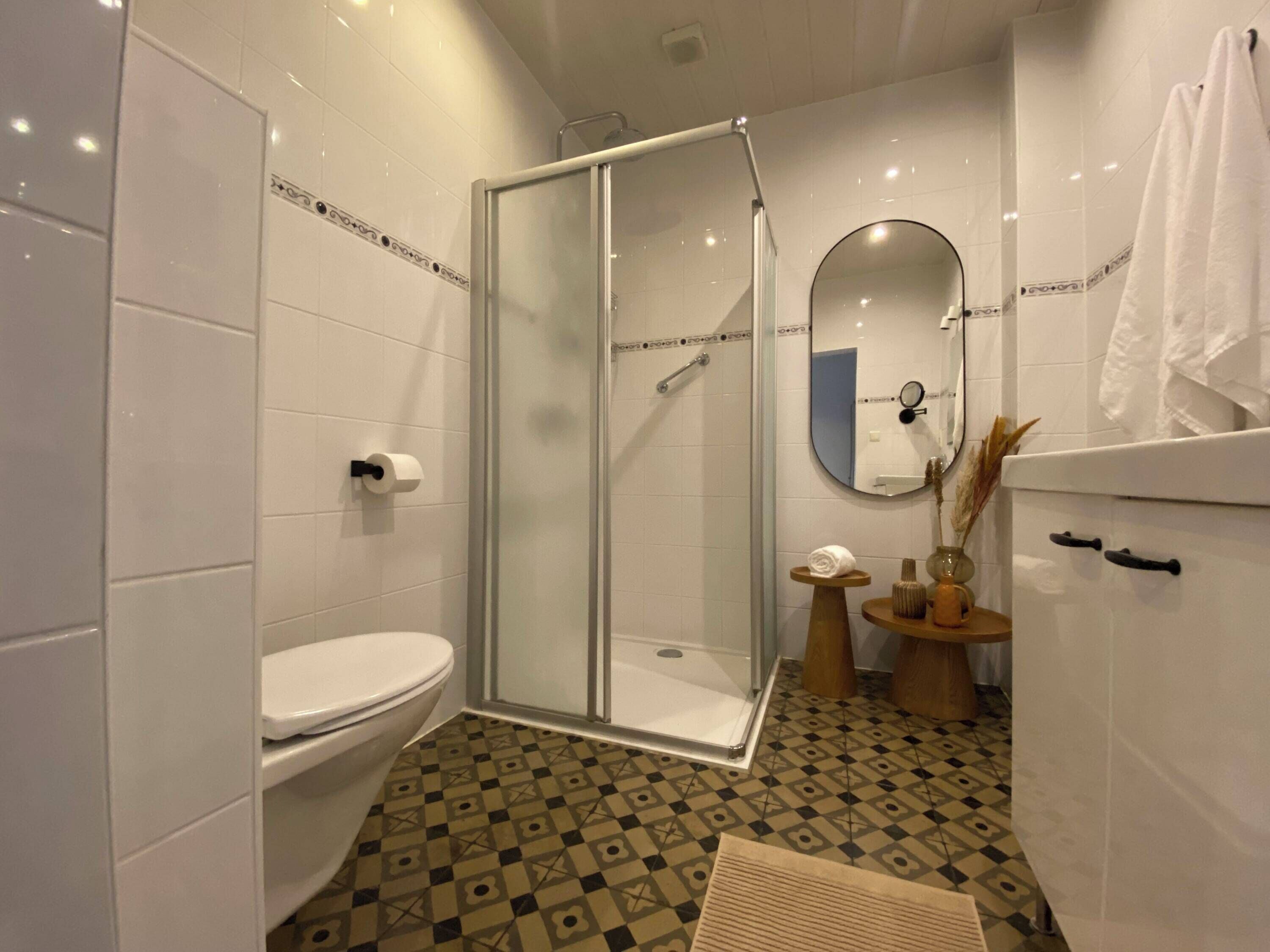 A modern bathroom with a glass-enclosed shower, a toilet, a sink with a mirror, and patterned floor tiles.