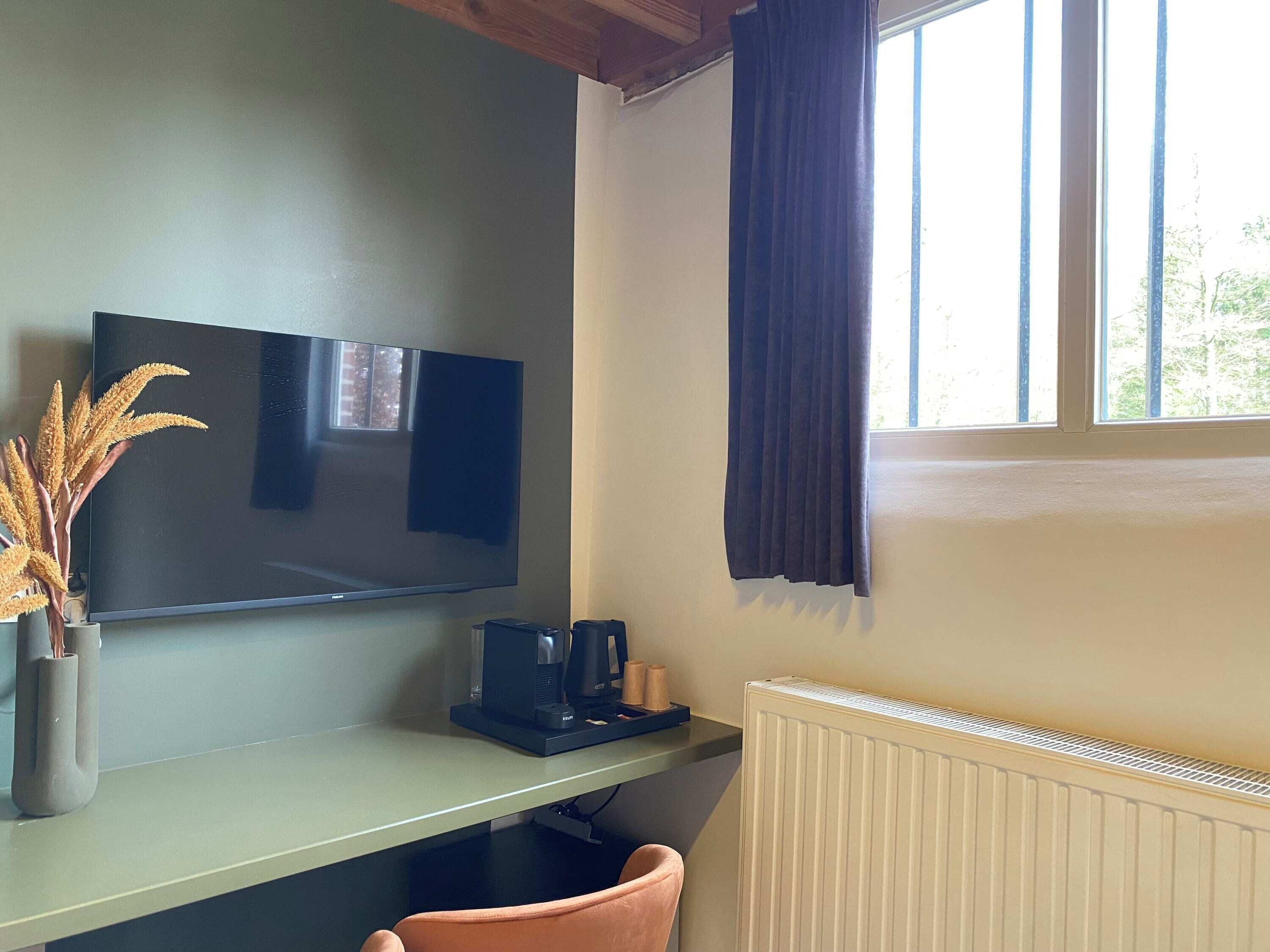 A room with a flat-screen TV mounted on the wall, a coffee maker on a table, a radiator, and a window with curtains.