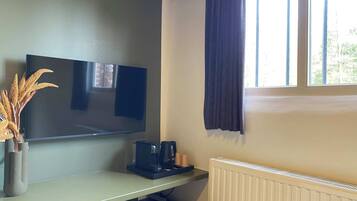 A room with a flat-screen TV mounted on the wall, a coffee maker on a table, a radiator, and a window with curtains.