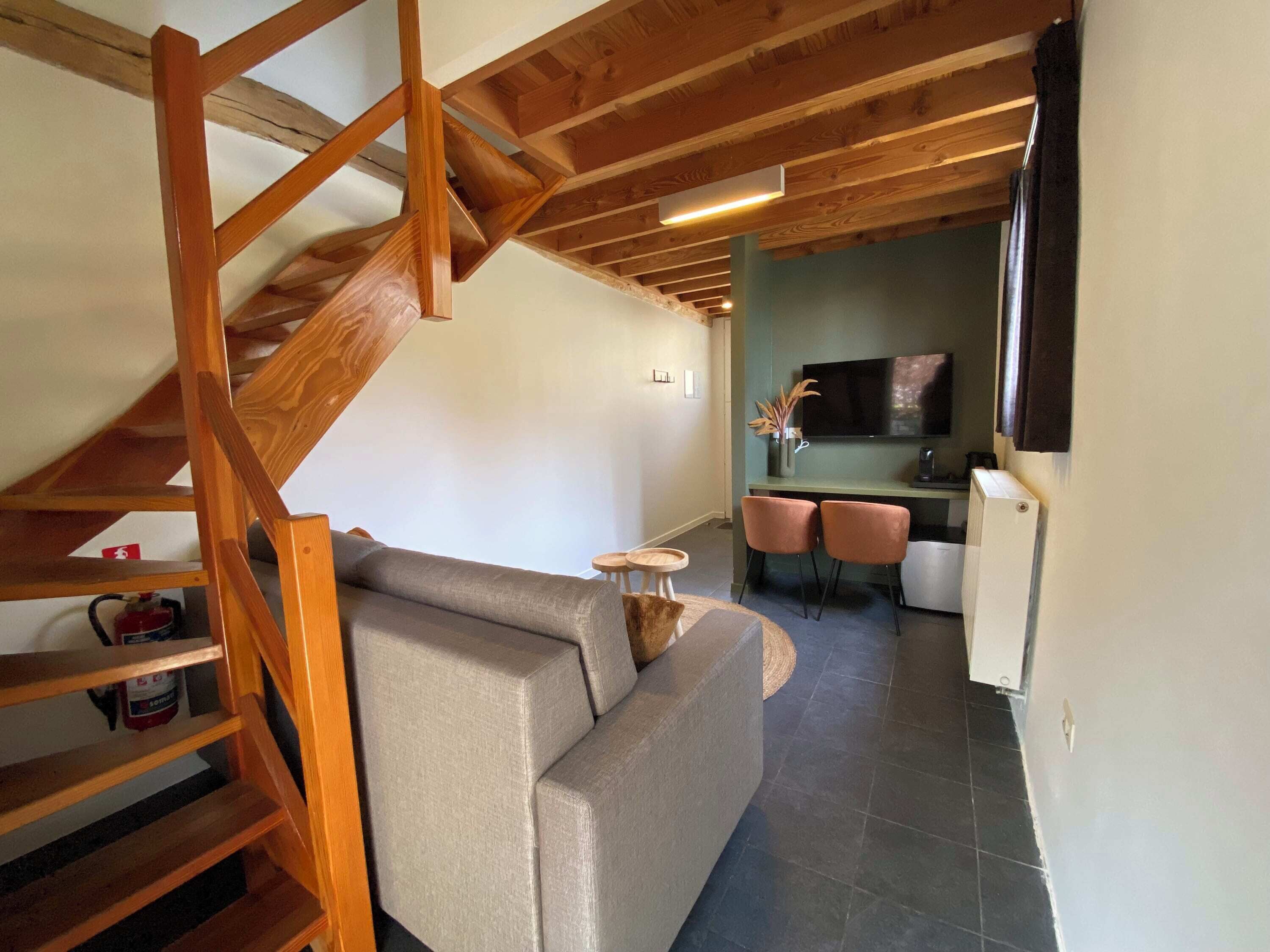 Un espace de vie moderne avec un escalier en bois, un canapé gris, une petite table ronde et un coin cuisine équipé d’une télévision et de deux chaises.