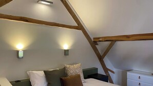 A bedroom with a sloped ceiling, wooden beams, a bed with pillows, a nightstand, and wall-mounted lights. - A1. De Fries: duplexsuite with terrace (Oudsbergen)