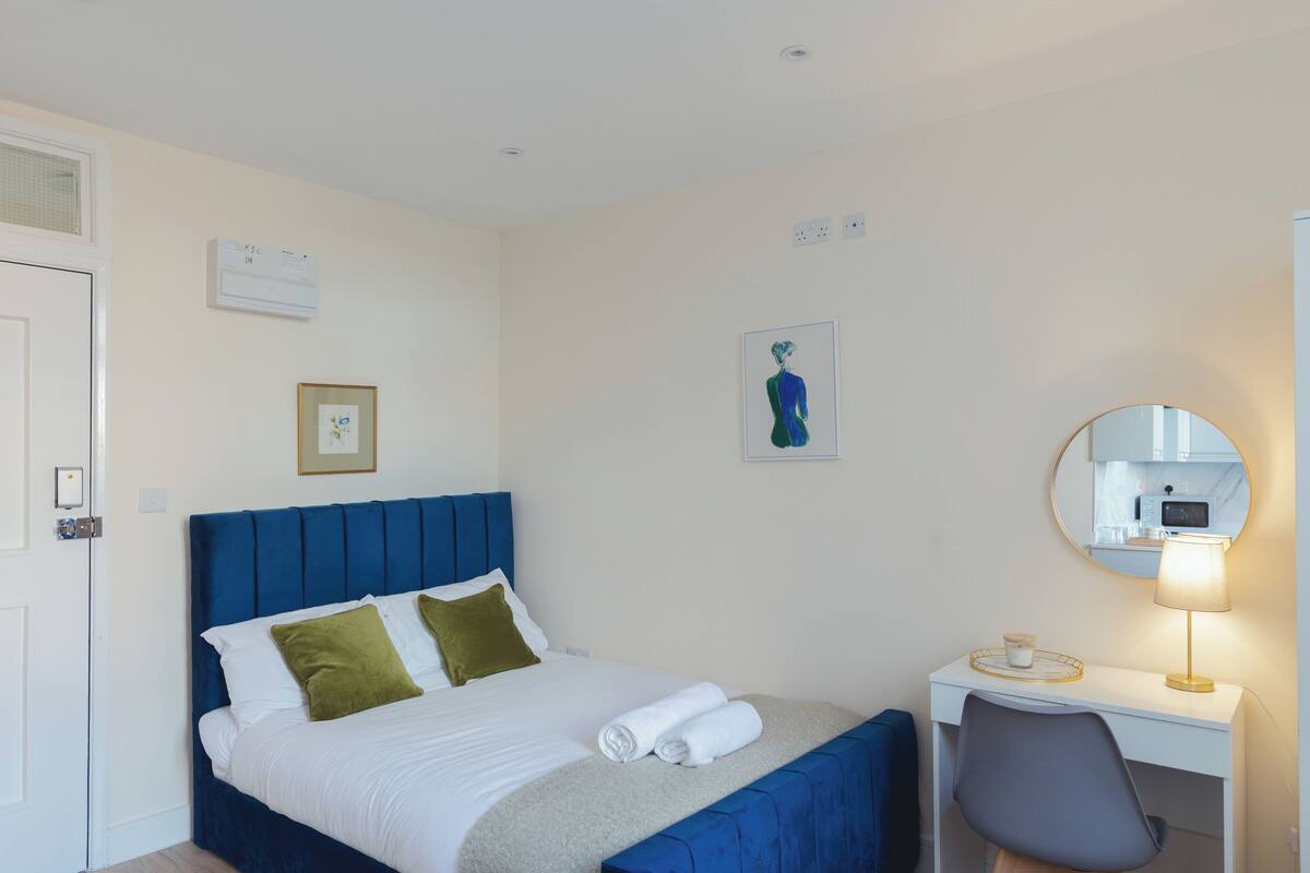 A hotel room with a bed, a desk, a chair, a round mirror, and a wall-mounted air conditioner.