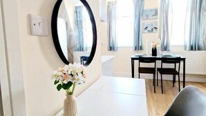 Une pièce moderne avec un bureau blanc, un miroir rond, un vase avec des fleurs et un coin repas comprenant une table et des chaises.