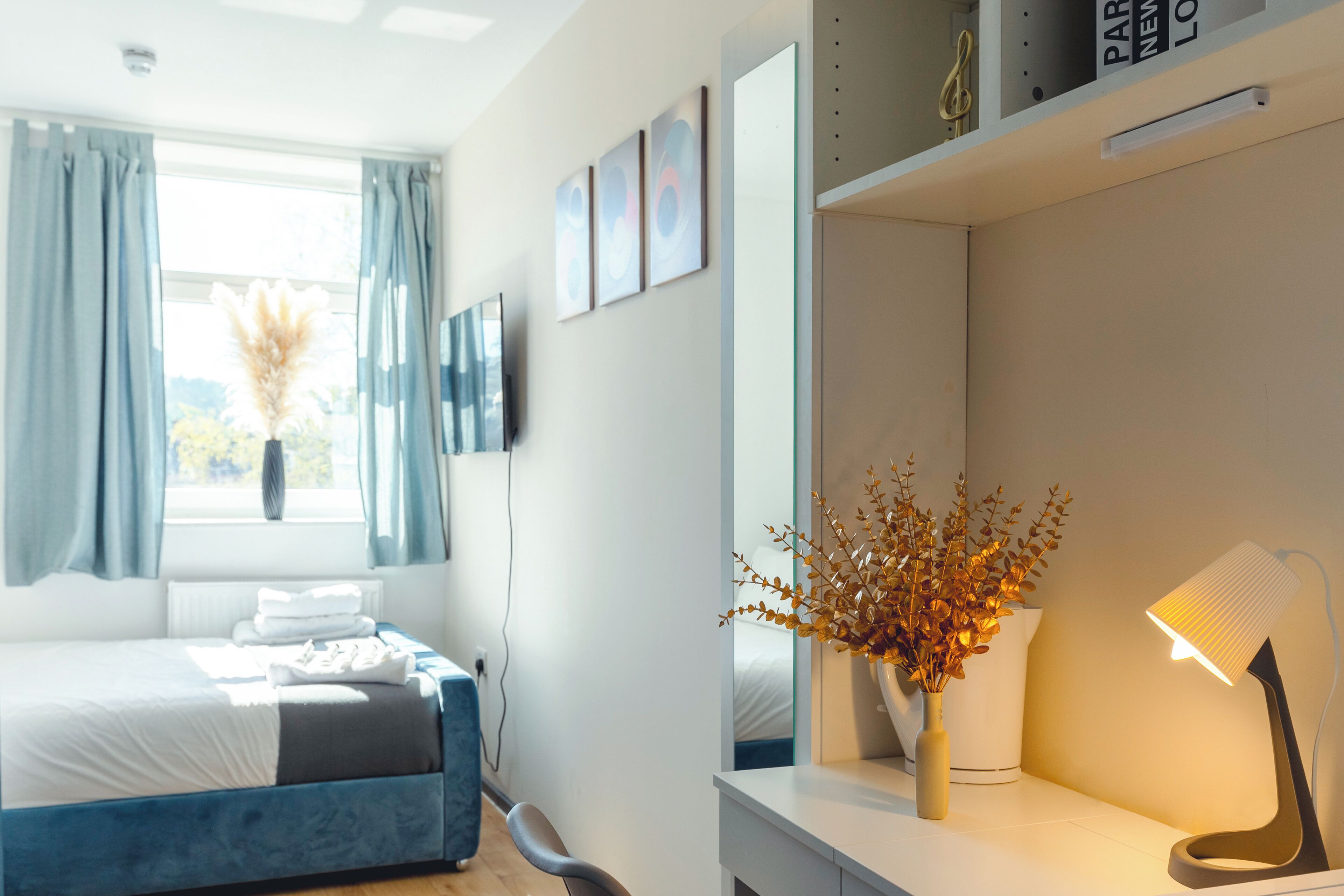 A modern hotel room with a bed, a desk with a lamp, a vase with dried flowers, and a window with curtains.