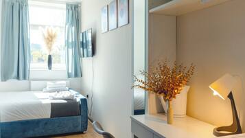 A modern hotel room with a bed, a desk with a lamp, a vase with dried flowers, and a window with curtains.