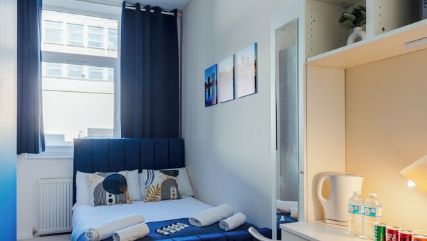 Une chambre d’hôtel bien rangée, avec un lit, un bureau, une bouilloire et une étagère où sont posées des bouteilles d’eau et des canettes de soda.