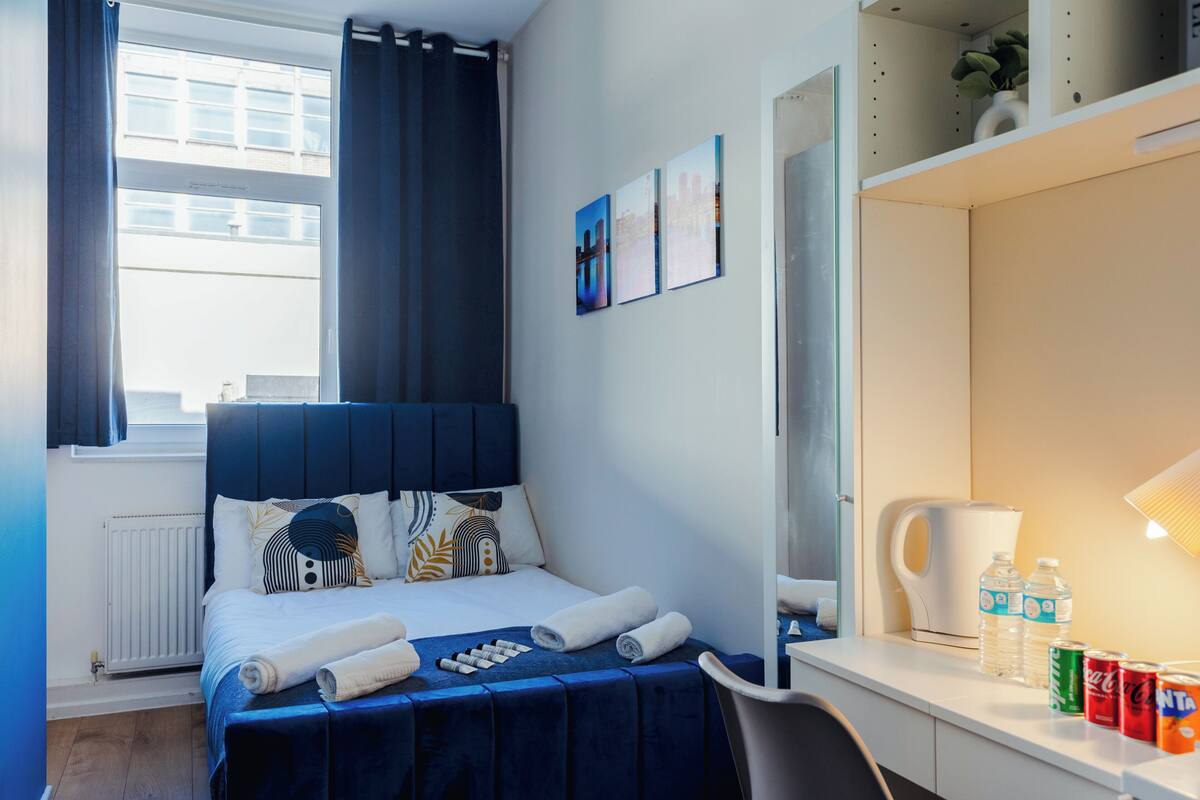 A neatly arranged hotel room with a bed, a desk, a kettle, and a shelf with bottled water and soda cans.