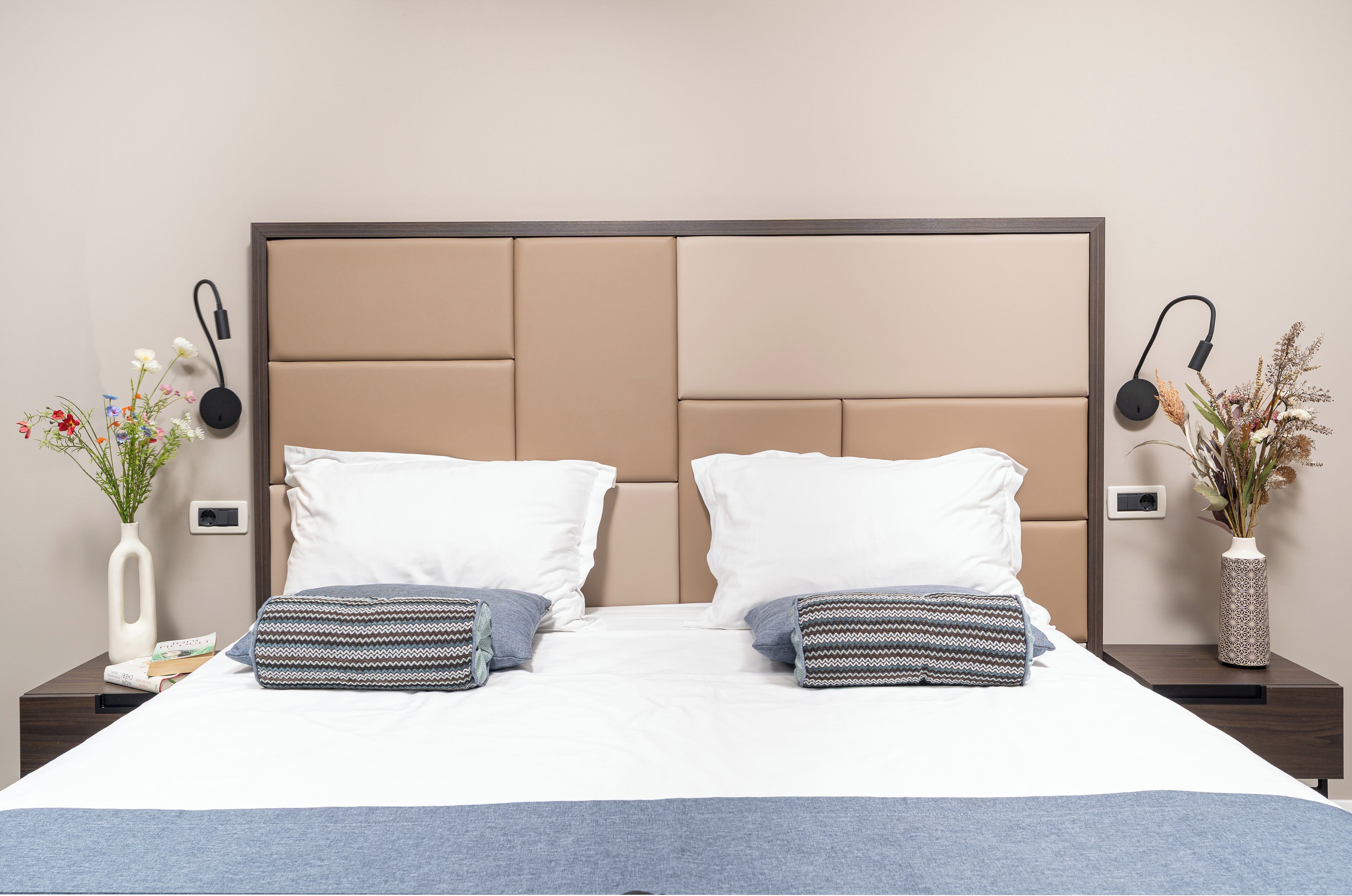 A neatly made bed with two pillows, a bedside table with a vase of flowers, and a wall-mounted charging station.