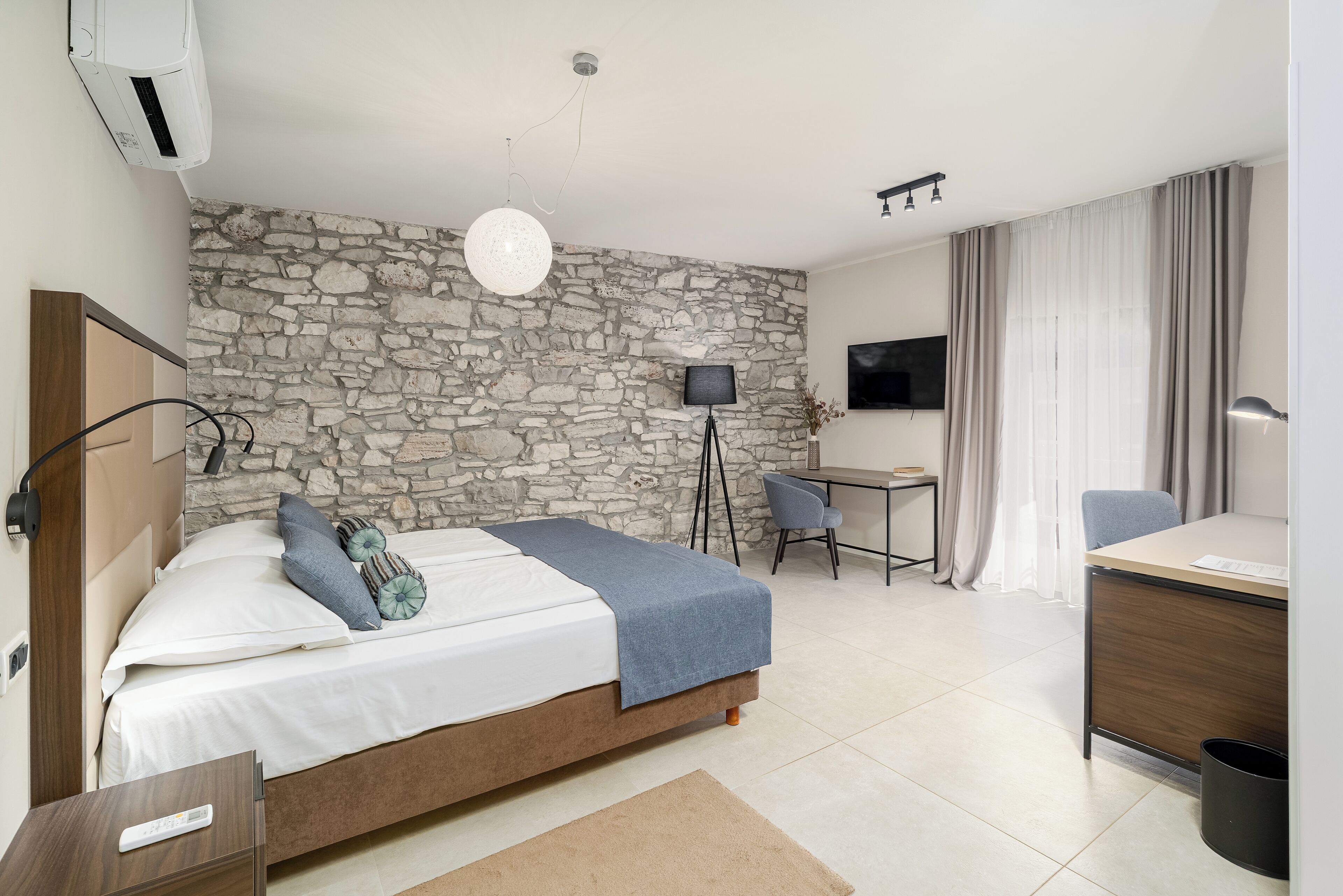 Quarto de hotel moderno com parede de pedra, cama com manta azul, escrivaninha com cadeira, televisão e janela com cortinas.