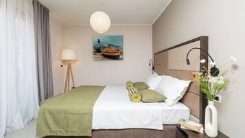 A neatly made bed with a green blanket, a bedside table with a vase of flowers, and a painting of boats on the wall.