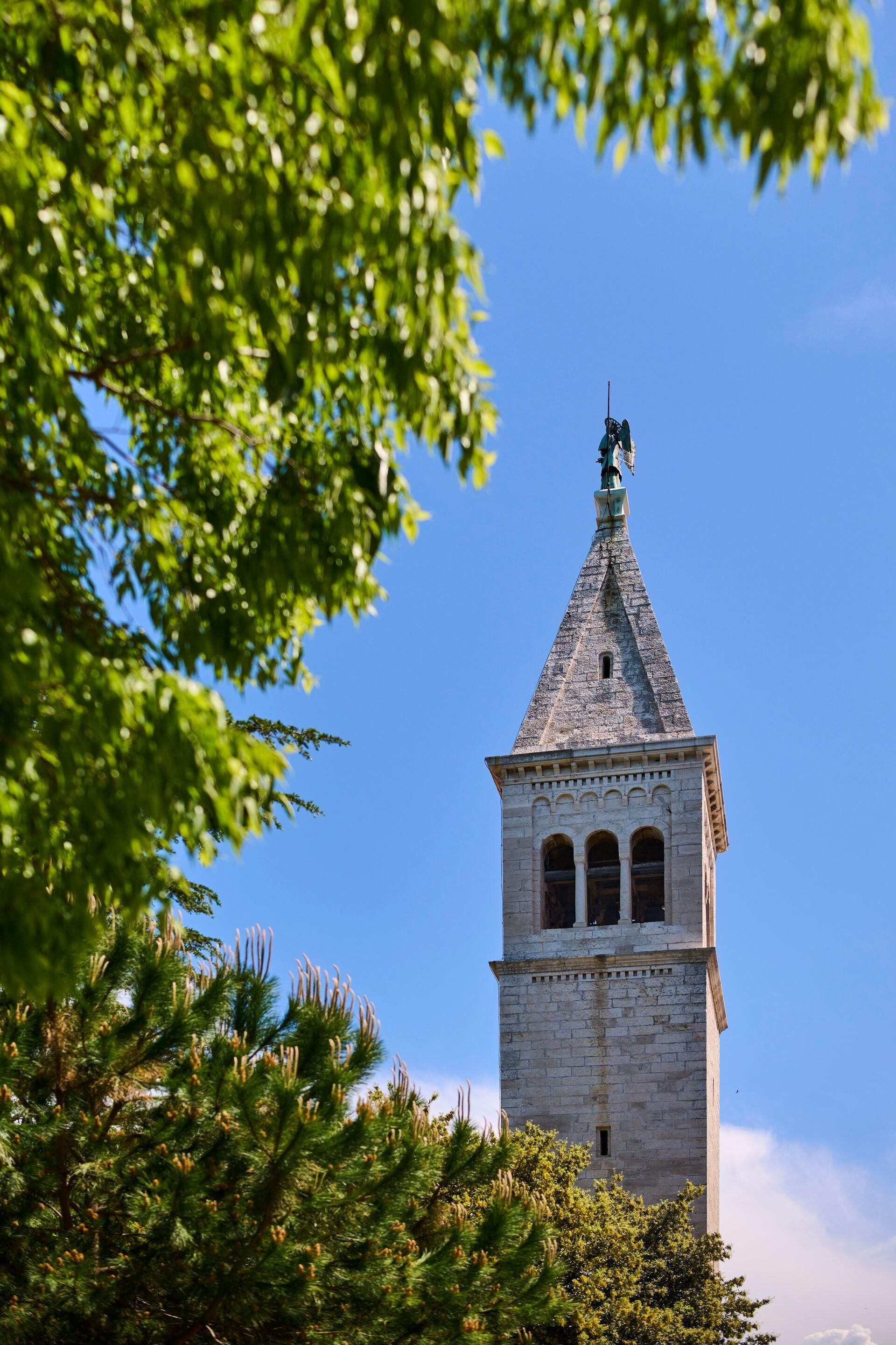Ein Turm mit spitzer Kuppel und einer Statue auf der Spitze, umgeben von Bäumen.