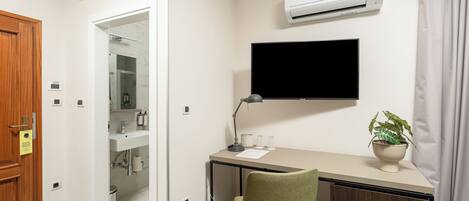A room with a desk, chair, TV, air conditioning unit, and a bathroom visible through an open door.