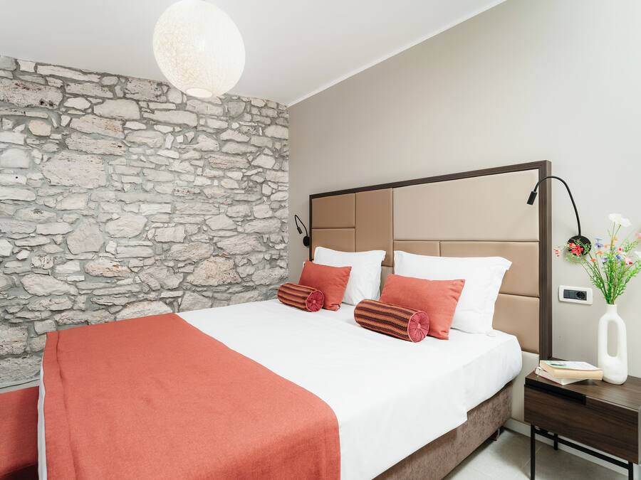 A bedroom with a stone wall, a bed with a wooden headboard, a nightstand, a lamp, and a vase with flowers.