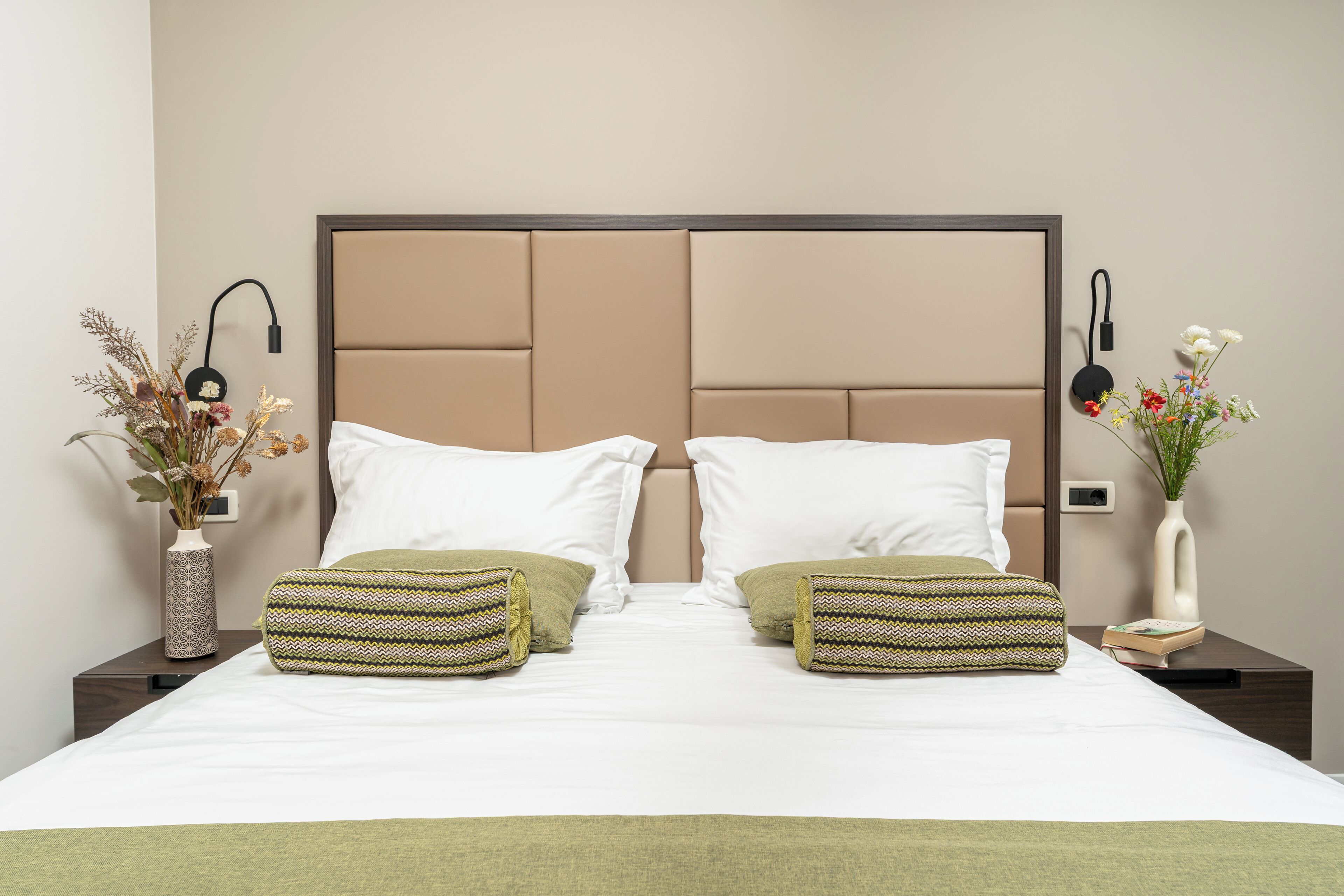 A neatly made bed with white linens, two green patterned pillows, and a beige headboard. There are two bedside tables, each with a vase of dried flowers and a book.