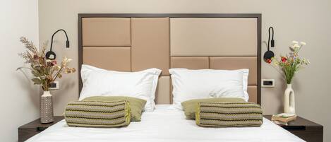 A neatly made bed with white linens, two green patterned pillows, and a beige headboard. There are two bedside tables, each with a vase of dried flowers and a book.
