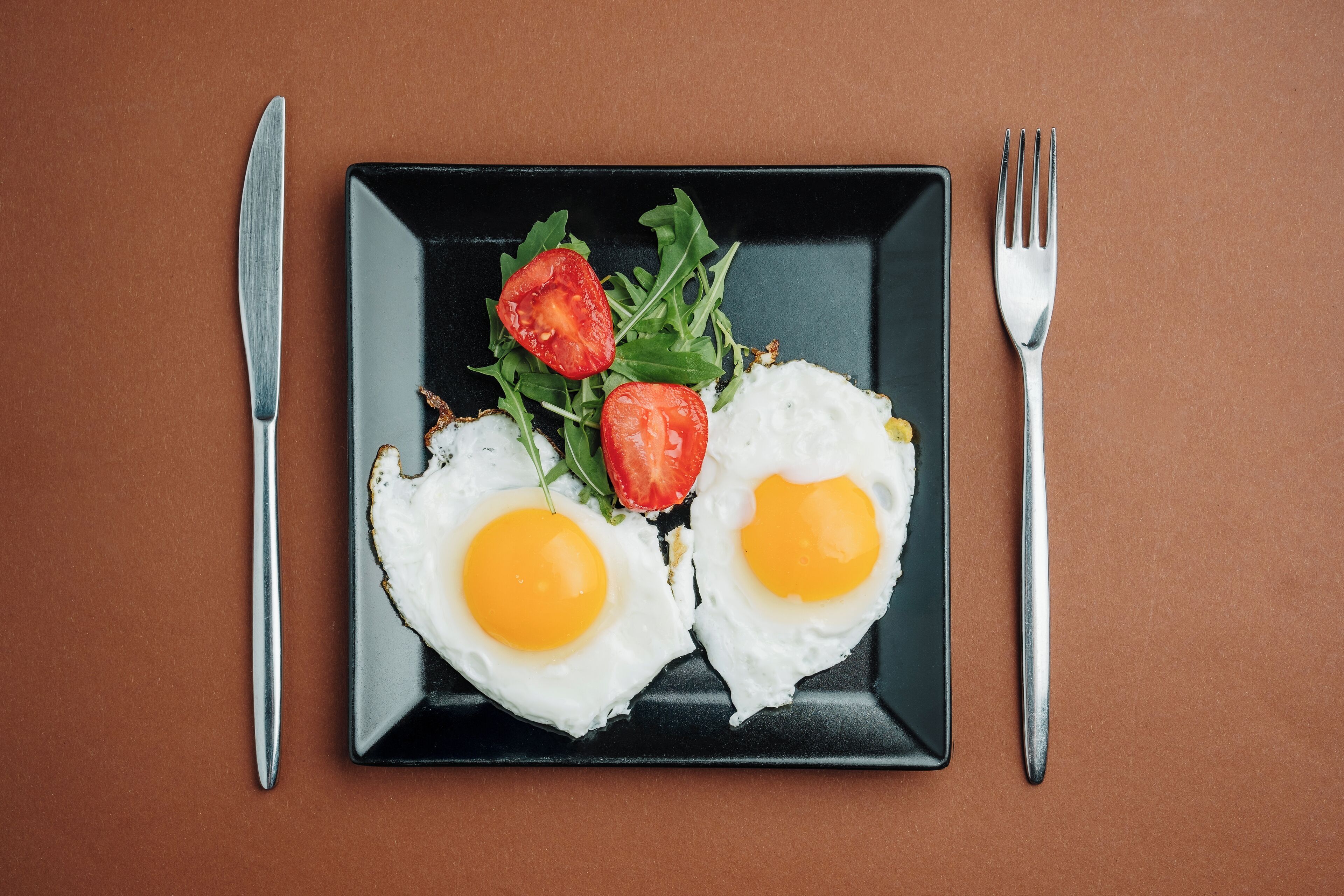 Une assiette noire sur laquelle on voit deux œufs au plat, des tomates cerises et de la roquette, le tout posé sur une surface marron.
