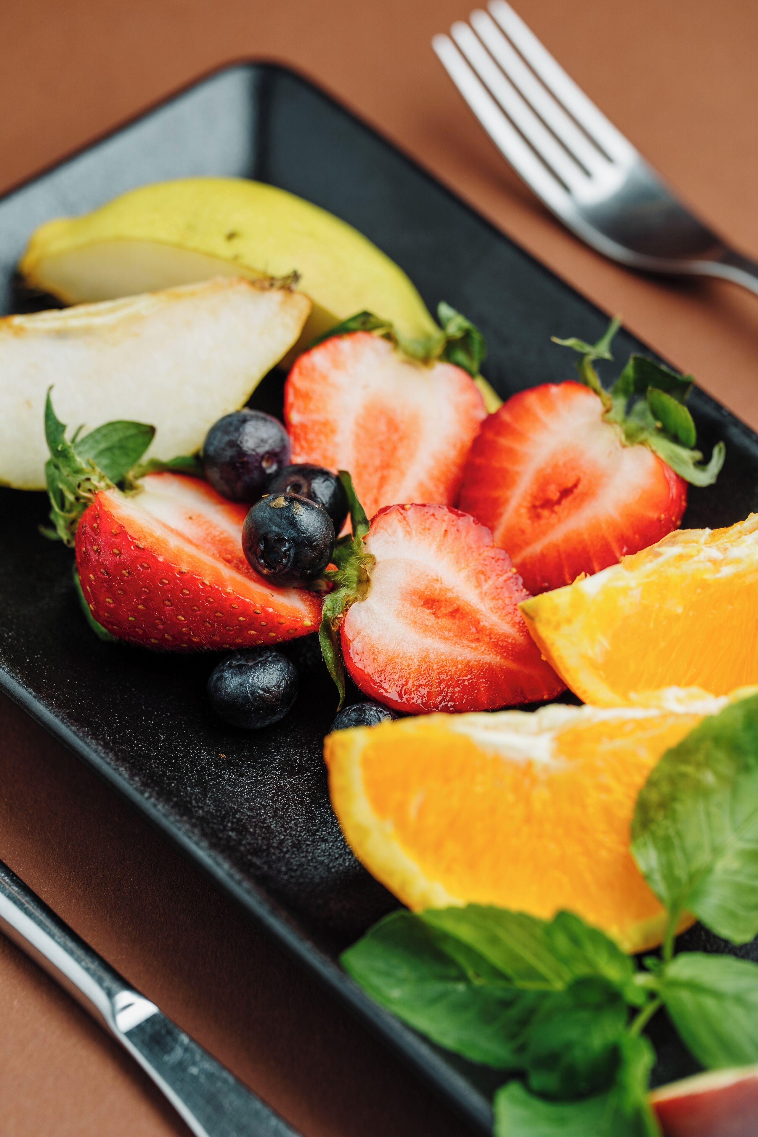 Ein schwarzes, rechteckiges Tablett mit Obst, darunter Erdbeeren, Blaubeeren und eine Orangenscheibe, dazu Minzblätter.