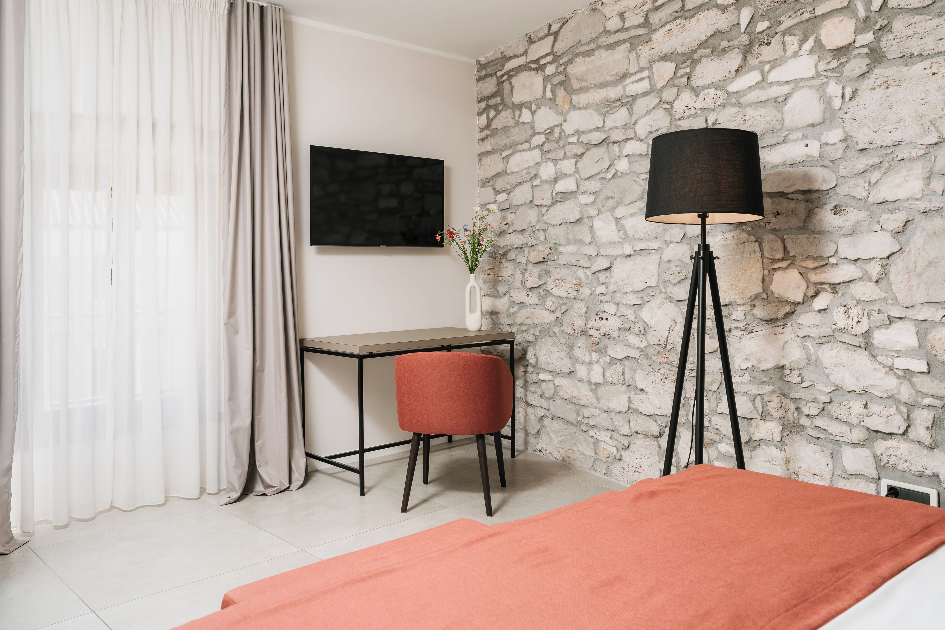 Une pièce avec un mur en pierre, un bureau avec une chaise, un lit recouvert d’une couverture orange, une lampe sur pied et un téléviseur fixé au mur.