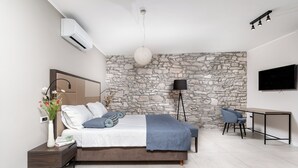 A modern bedroom with a stone wall, a bed with a blue blanket, a bedside table with a vase of flowers, a desk with a chair, and a mounted TV. - Al Porto Suites (Novigrad)