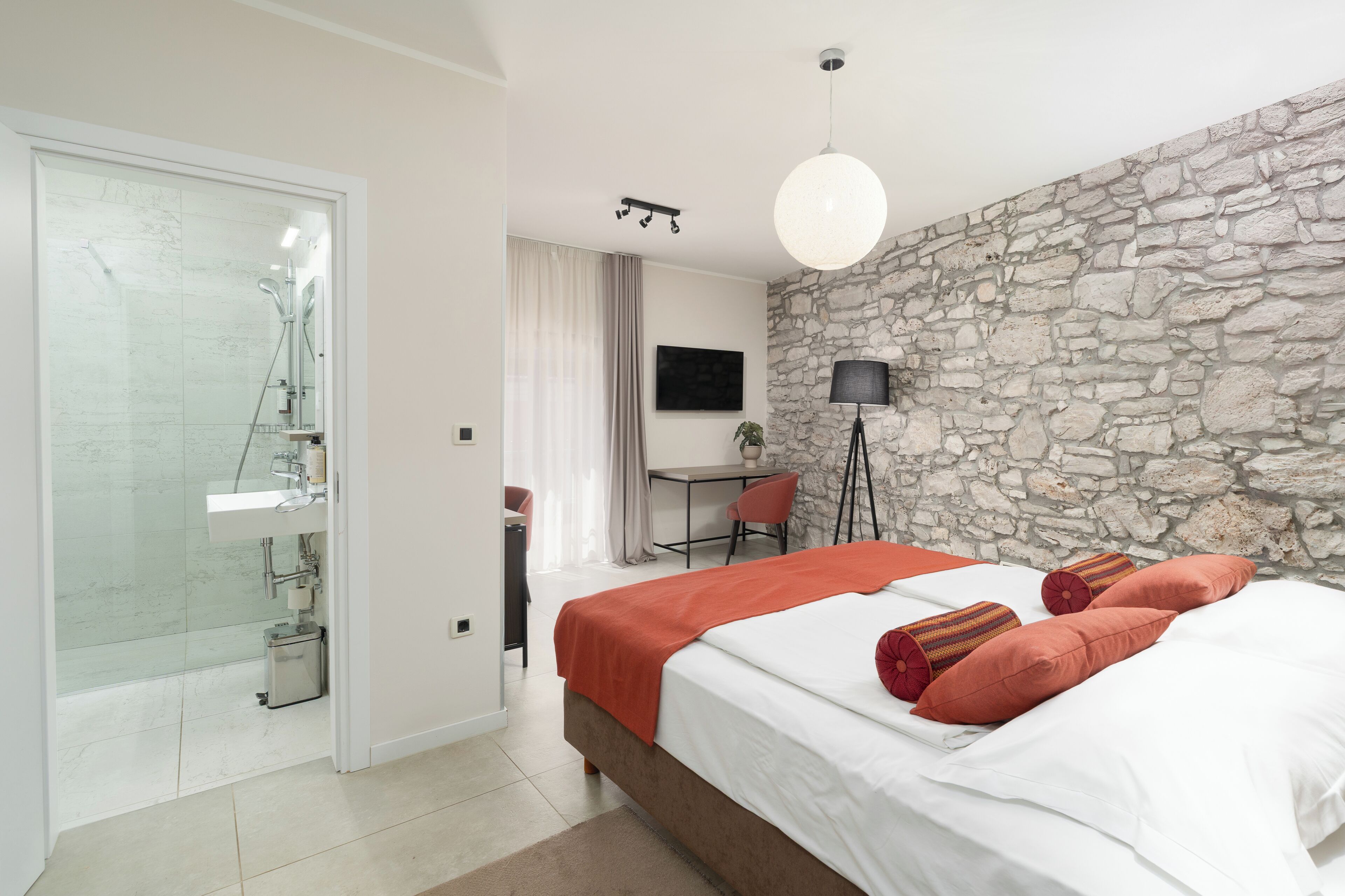 A modern bedroom with a stone wall, a bed with red pillows, a desk with a chair, and a shower area.
