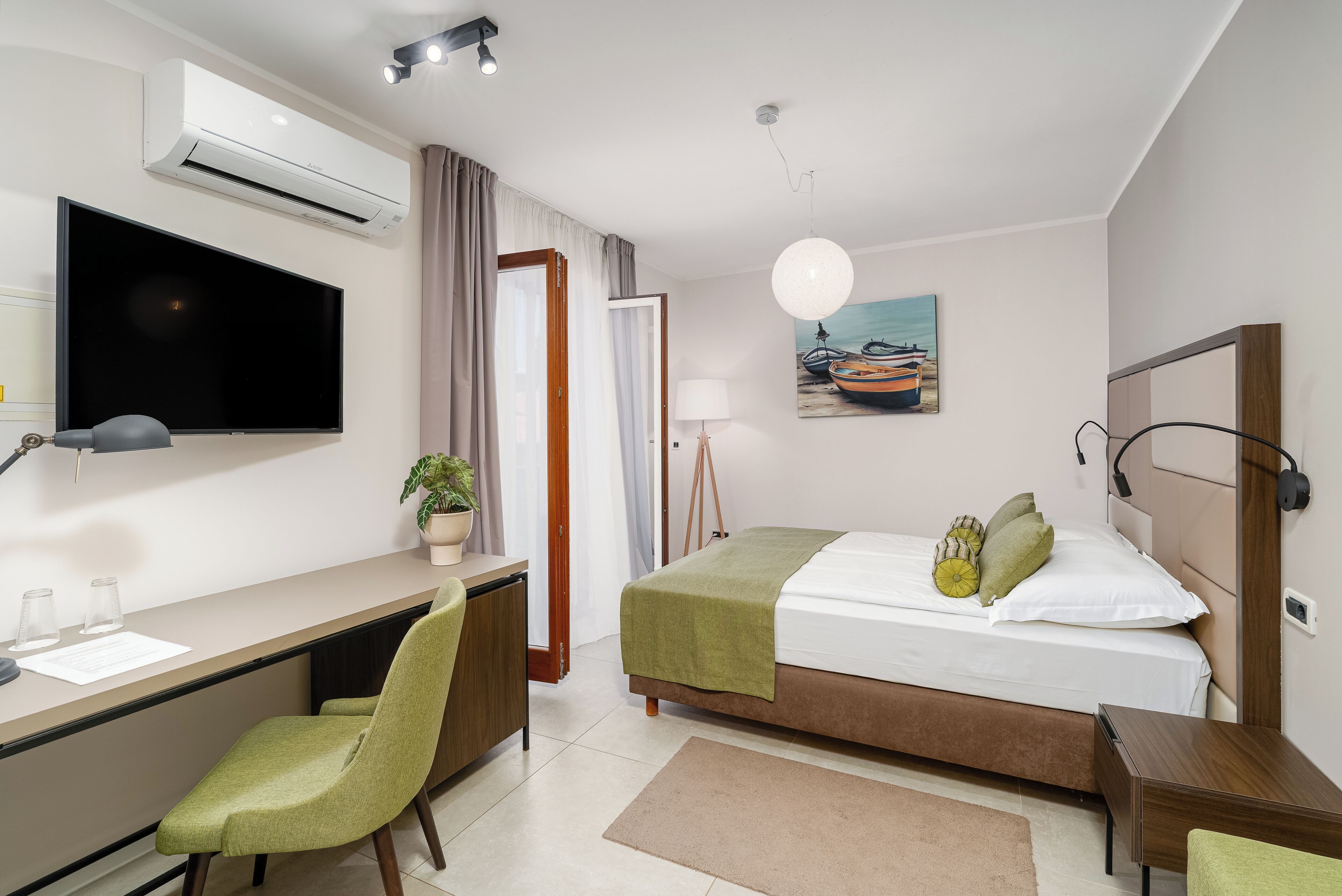 A modern hotel room with a flat-screen TV, a desk with a chair, a bed with a green blanket, a bedside table, and a wall-mounted lamp.