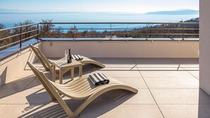 Un balcon avec deux fauteuils de jardin en bois, une bouteille de vin et un verre. Le balcon offre une vue sur une étendue d’eau et sur des montagnes lointaines.